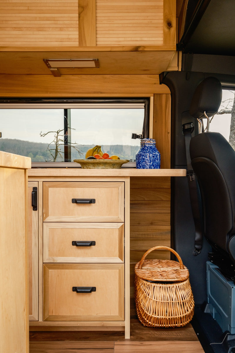 RV rental interior: Warm wooden kitchen with drawers, fruit bowl, scenic window view, passenger seat, and wicker basket. — photo 4