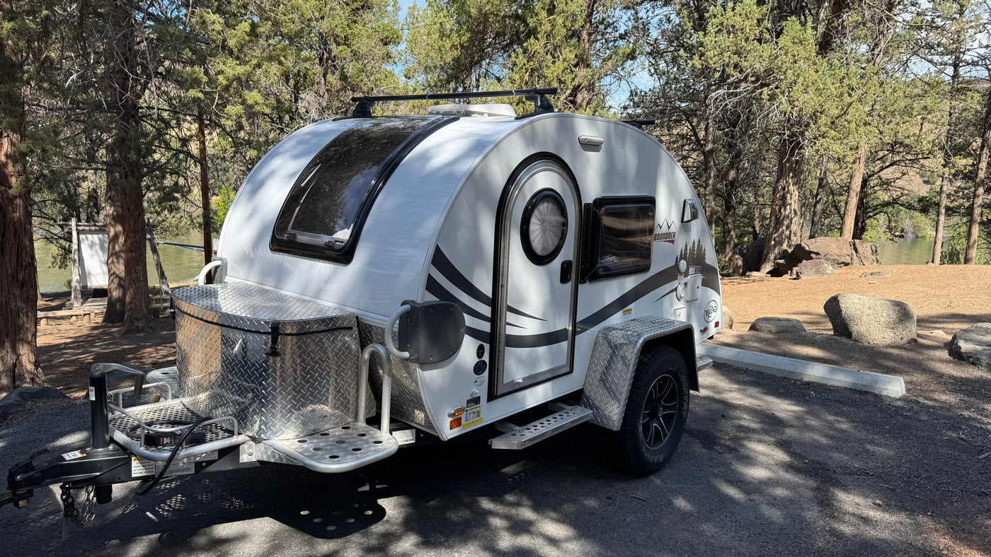 White nuCamp T@G Boondock teardrop **rv rental** parked at a wooded campsite, ideal for nature getaways by the water.