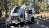 White nuCamp T@G Boondock teardrop **rv rental** parked at a wooded campsite, ideal for nature getaways by the water.