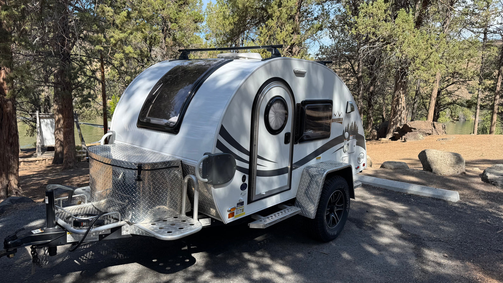 White nuCamp T@G Boondock teardrop **rv rental** parked at a wooded campsite, ideal for nature getaways by the water.