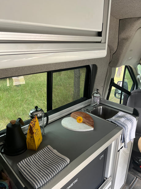 Inside an RV rental: a well-equipped kitchenette with coffee, sink, Dometic fridge, and a green outdoor view.