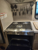 An RV rental kitchen features a stainless steel Dometic stove and oven with an open door, and cooking utensils on a magnetic strip.