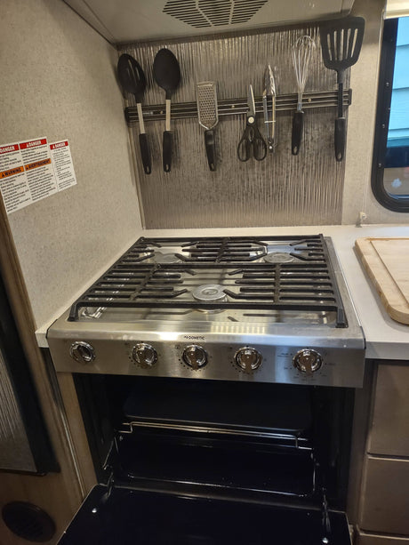 An RV rental kitchen features a stainless steel Dometic stove and oven with an open door, and cooking utensils on a magnetic strip.