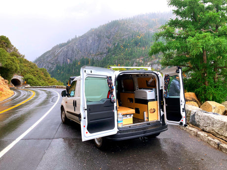 RV rental camper van parked on a wet mountain road, back doors open to show its cozy interior amidst mountains and trees.
