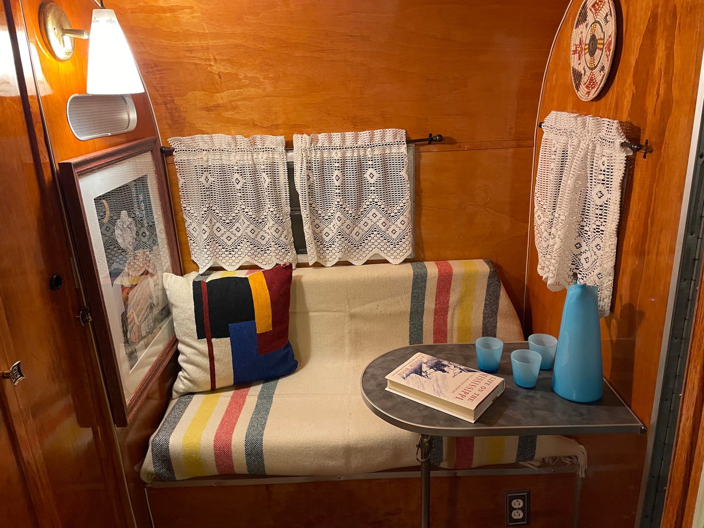 Cozy rv rental interior featuring polished wood, a striped bench, lace curtains, and a small table with a book and blue glass decor.