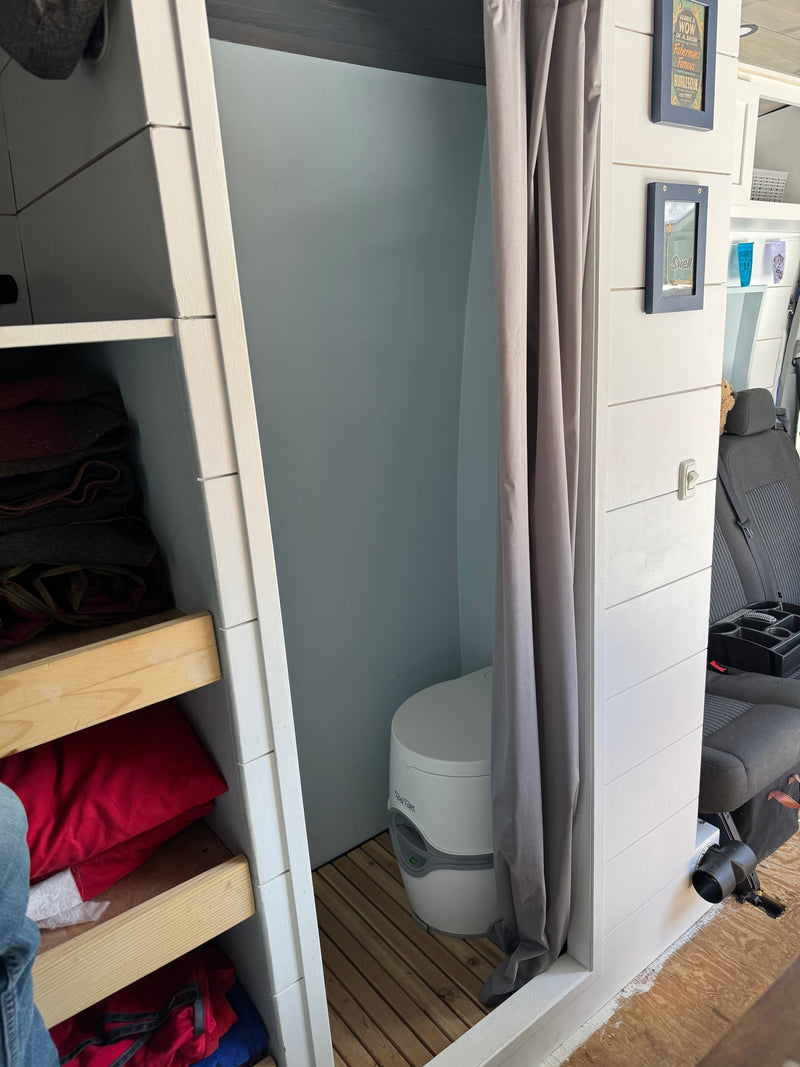 Explore this RV rental's camper van interior. Features a compact bathroom with a portable toilet behind a curtain, next to storage shelves. — photo 6