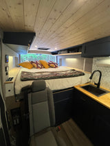 Cozy RV rental camper van interior. Features a raised bed looking into trees, wood ceiling, and compact kitchen.