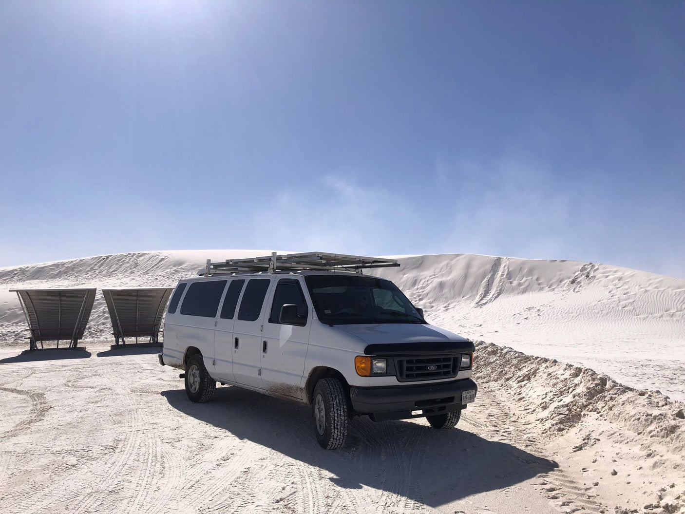 White Ford van (RV rental) on sandy terrain near large white dunes under blue sky. Two shelters behind. Ready for desert adventures.