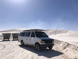 White Ford van (RV rental) on sandy terrain near large white dunes under blue sky. Two shelters behind. Ready for desert adventures.