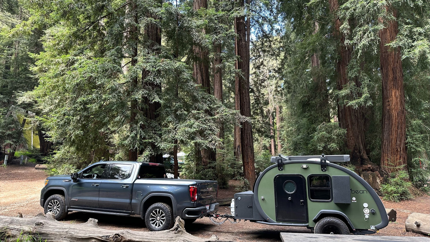 Discover the perfect RV rental: a GMC truck towing a green teardrop camper amidst towering redwoods.
