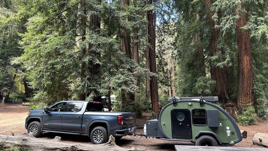 Discover the perfect RV rental: a GMC truck towing a green teardrop camper amidst towering redwoods.