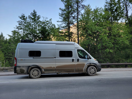 Mud-splattered grey Winnebago Solis camper van, likely an RV rental, on a mountain road surrounded by green trees.
