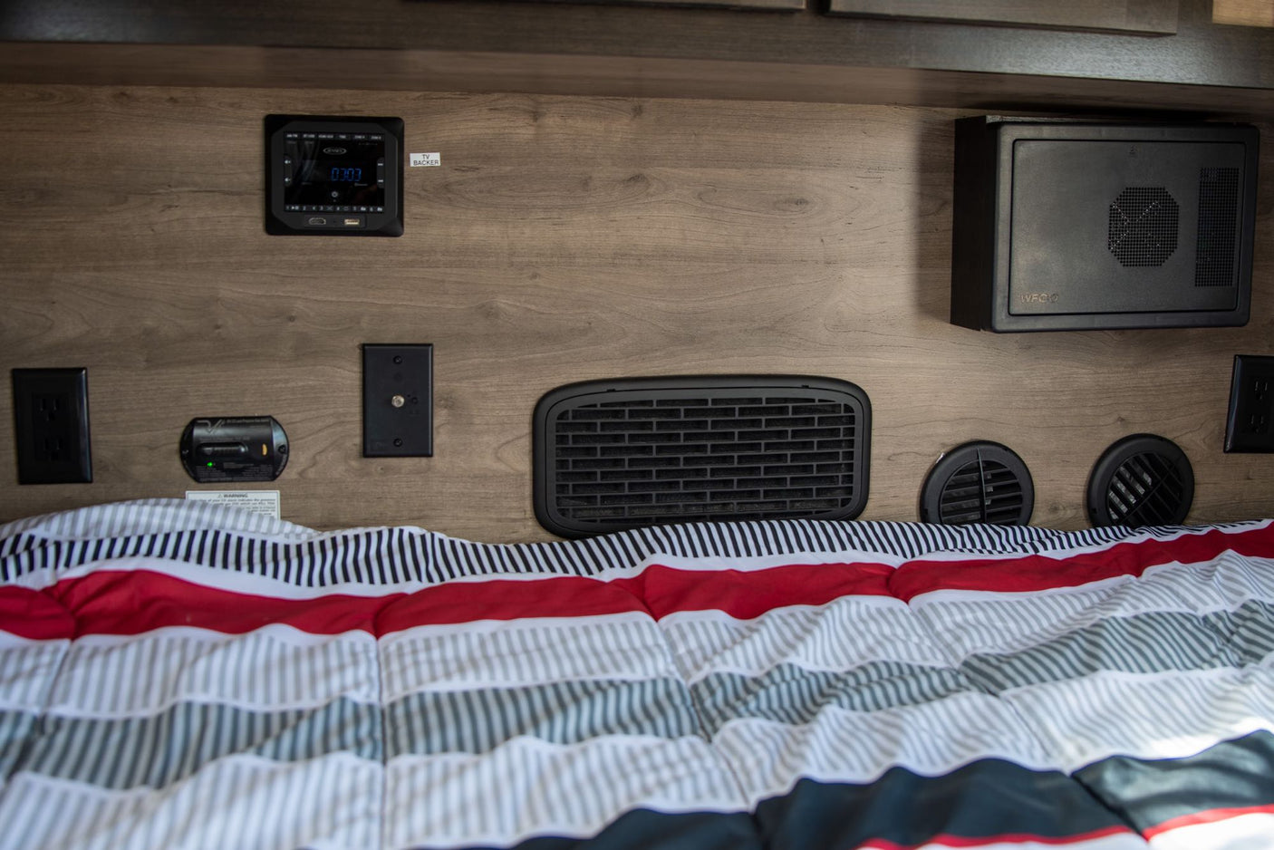 RV rental bedroom: Wall features Jensen audio, power outlets, CO detector, various vents, and a WFCO unit above a comfy bed.