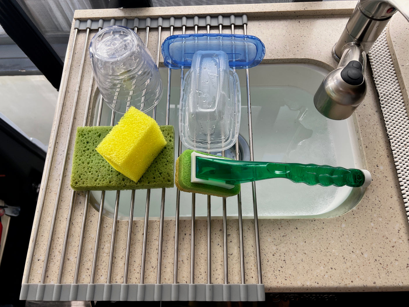 Compact RV rental kitchen sink with roll-up drying rack holding washed plastic cup, containers, and cleaning tools.