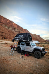 An adventure-ready RV rental Jeep with a rooftop tent set up in a vast desert landscape with red rock formations.