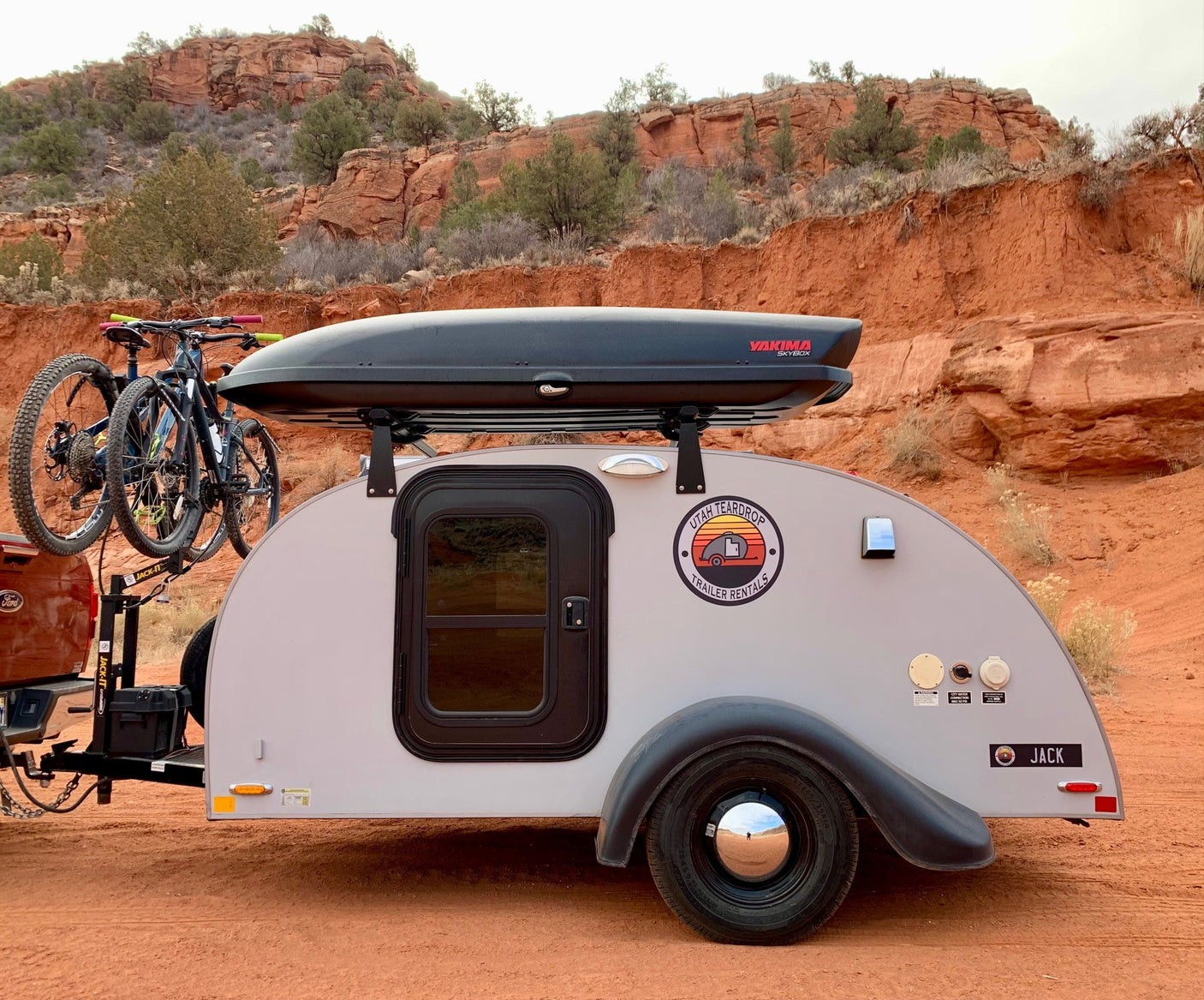 An RV rental teardrop trailer, "JACK," with bikes and a cargo box, parked on red earth by stunning rock formations, ready for adventure.