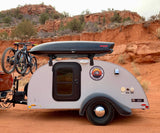 An RV rental teardrop trailer, "JACK," with bikes and a cargo box, parked on red earth by stunning rock formations, ready for adventure.