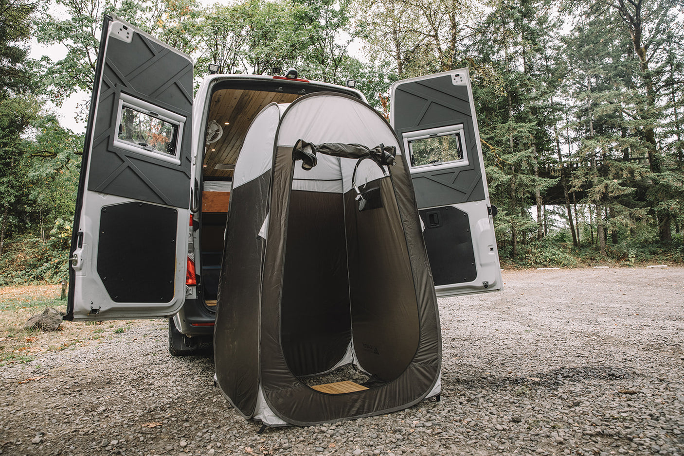 A white RV rental with open rear doors reveals its wooden interior, beside a pop-up portable shower tent for outdoor privacy.