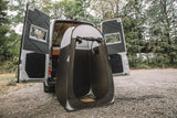 A white RV rental with open rear doors reveals its wooden interior, beside a pop-up portable shower tent for outdoor privacy.
