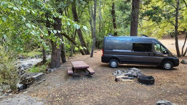 A gray rv rental camper van is parked by a forest stream with a picnic table and fire pit, ideal for nature camping. — photo 10
