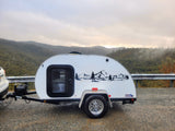 Scenic rv rental: White teardrop trailer with mountain decal, parked on a gravel road overlooking foggy, forested mountains.