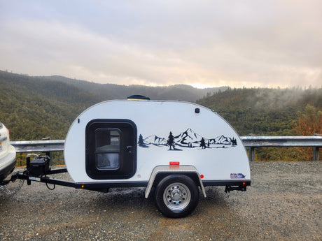 Scenic rv rental: White teardrop trailer with mountain decal, parked on a gravel road overlooking foggy, forested mountains.