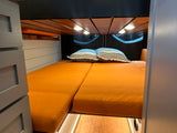 RV rental: View of a custom-built camper van bed, with vibrant orange cushions, striped pillows, and warm wooden ceiling lights.