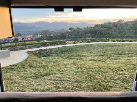 Panoramic view from an RV rental showcasing a grassy lawn, rolling hills, and distant mountains under a warm sky.