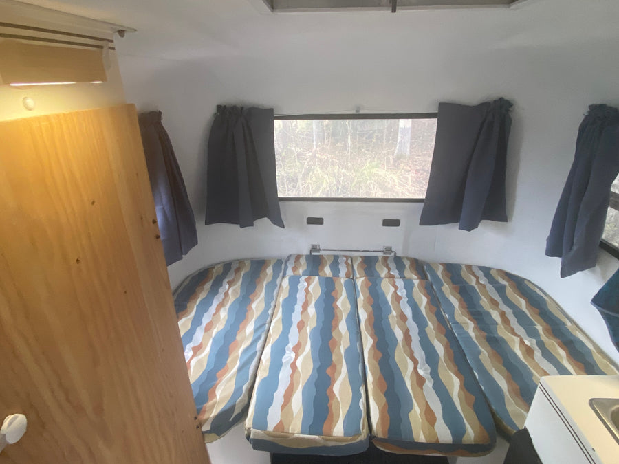 RV rental interior: A cozy sleeping area with colorful striped cushions, a large window showcasing nature, and blue curtains.