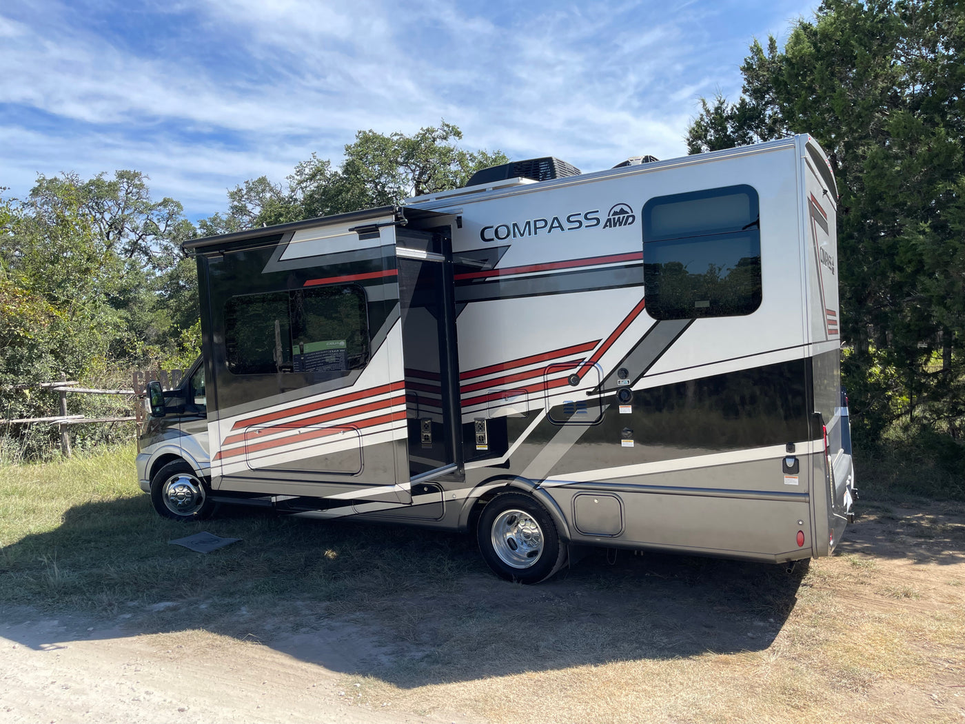 A sleek Compass AWD RV rental parked at a sunny campsite with its slide-out extended, surrounded by green trees and blue sky. — photo 11