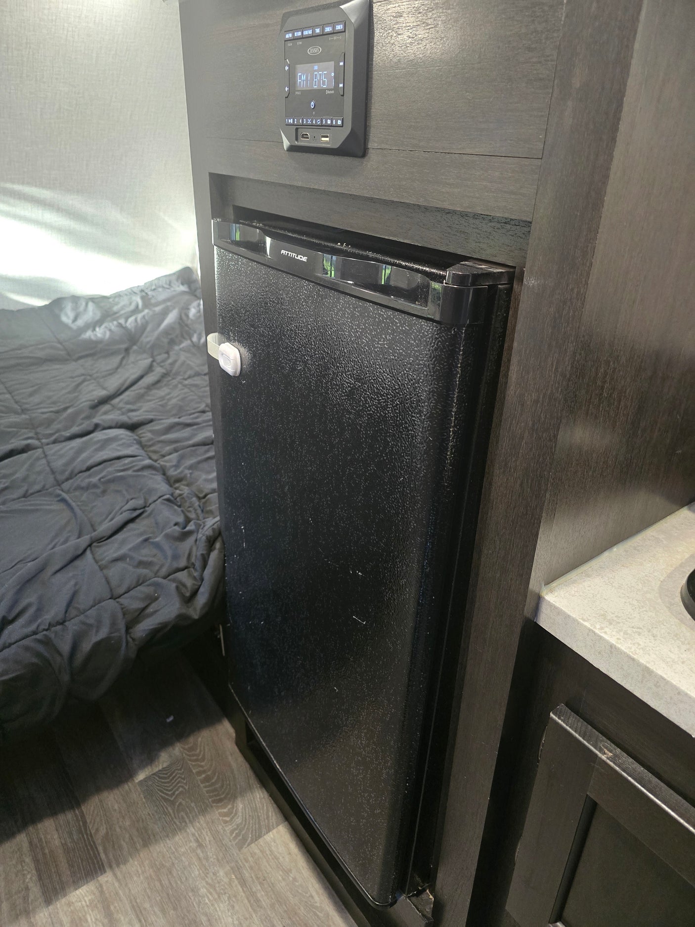 RV rental interior: Compact fridge with child lock, Jensen control panel above, and a grey quilted bed in the RV bedroom.