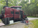 Red Winnebago Solis RV rental parked at a wooded campsite with pop-top open, chairs, and table set up outside.