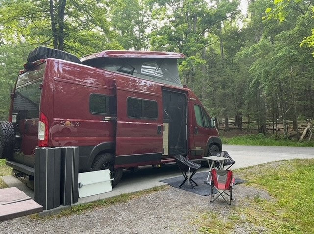 Red Winnebago Solis RV rental parked at a wooded campsite with pop-top open, chairs, and table set up outside. — photo 1