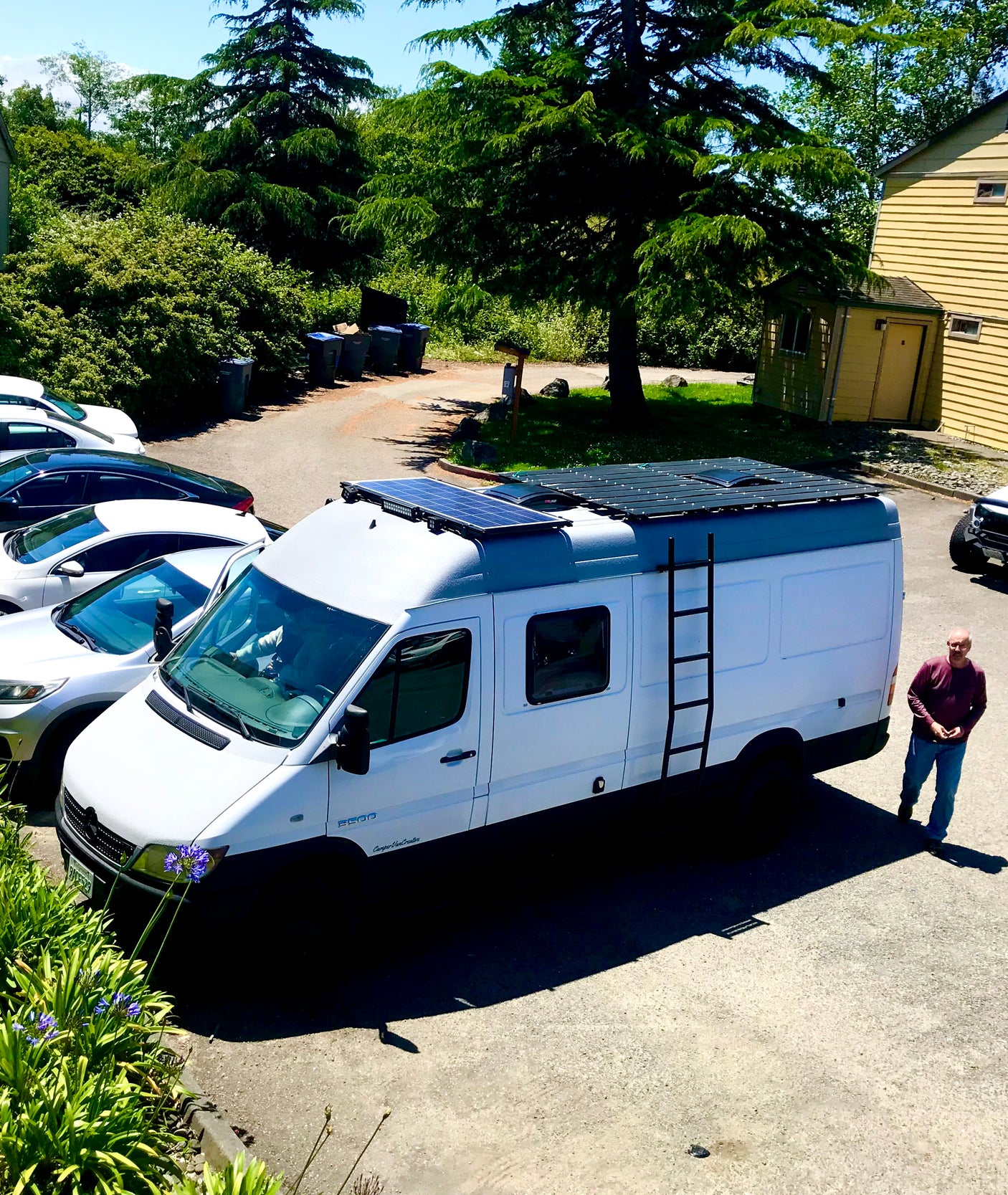 White Mercedes Sprinter camper van with solar panels on roof, ladder. Man by van. RV rental ready.