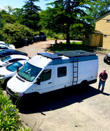 White Mercedes Sprinter camper van with solar panels on roof, ladder. Man by van. RV rental ready.