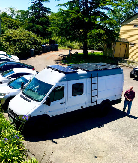 White Mercedes Sprinter camper van with solar panels on roof, ladder. Man by van. RV rental ready.