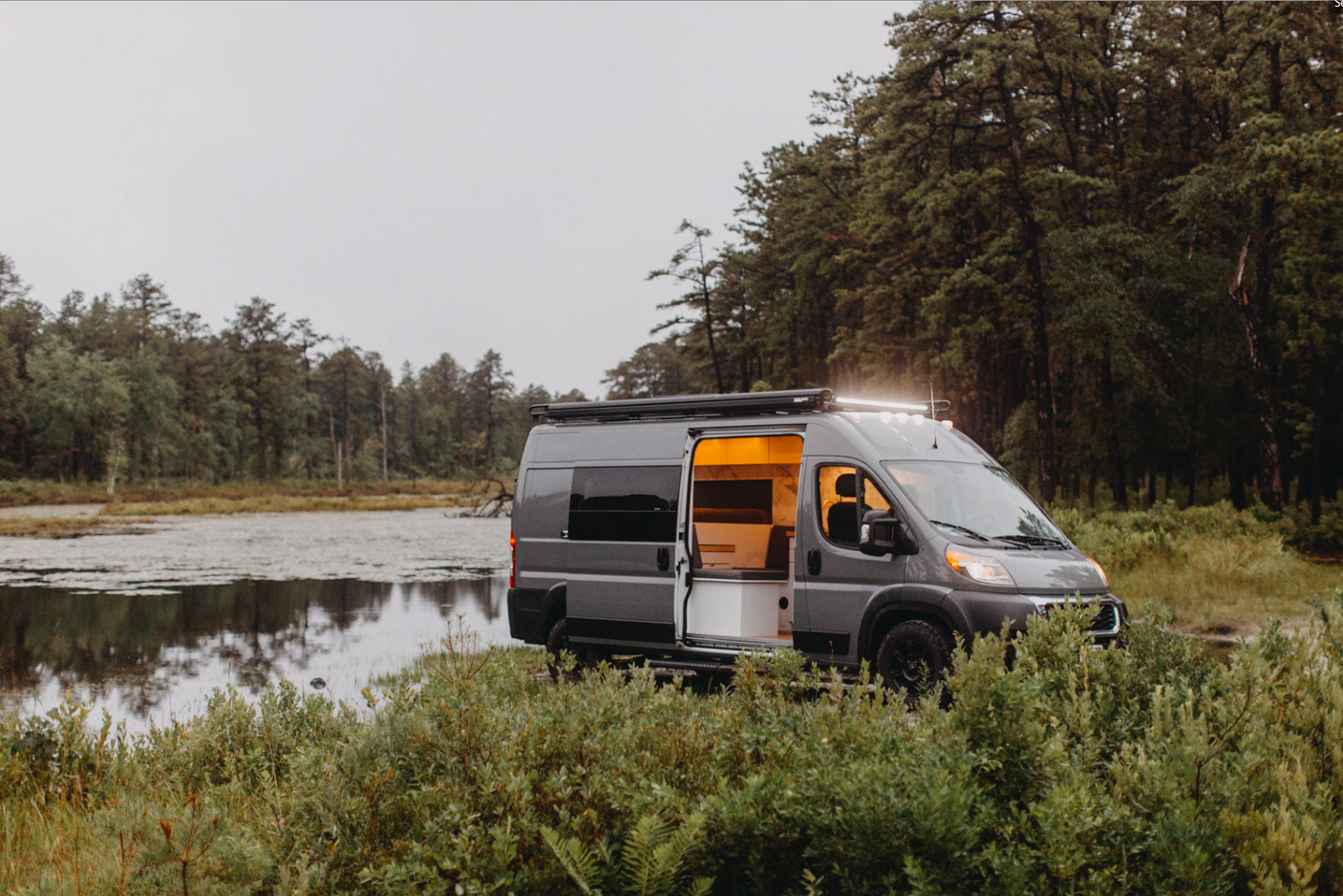 Grey camper van with illuminated interior and open door parked by a tranquil lake. Ideal rv rental for nature adventures.