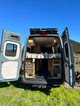 Open doors reveal a custom RV rental camper van interior with bed & storage, ready for adventure on grass.