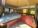 Inside this RV rental: a cozy bed with patterned bedding, woven ceiling, reddish cabinets, and a compact sink.