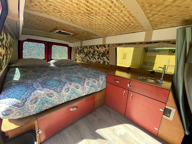 Inside this RV rental: a cozy bed with patterned bedding, woven ceiling, reddish cabinets, and a compact sink.