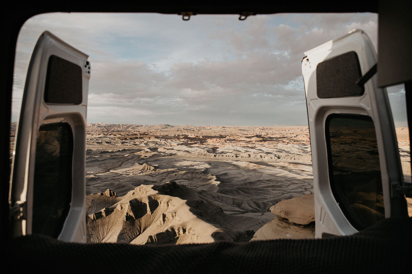 Open RV rental doors frame a stunning desert vista. Textured badlands and distant mesas stretch under a vast, cloudy sky.