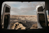 Open RV rental doors frame a stunning desert vista. Textured badlands and distant mesas stretch under a vast, cloudy sky.
