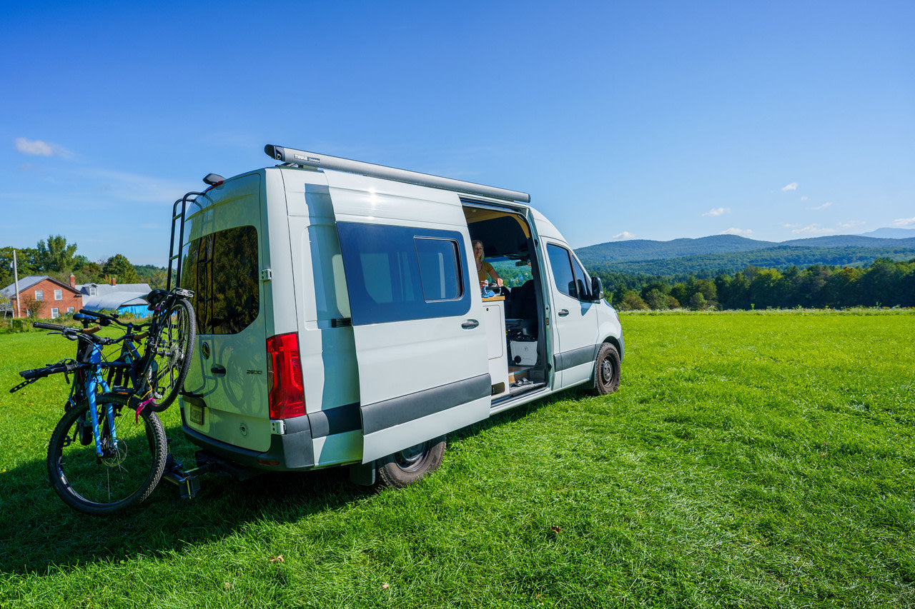 Explore nature with an RV rental: A white camper van with two bikes on the back is parked in a green field, mountains beyond.