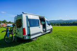 Explore nature with an RV rental: A white camper van with two bikes on the back is parked in a green field, mountains beyond.