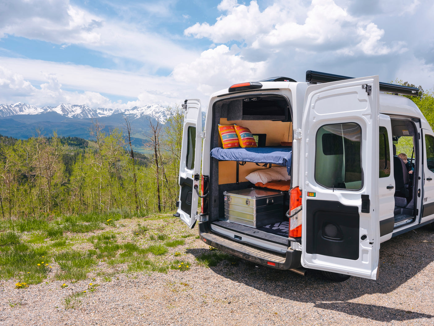 An RV rental, a white camper van with open back doors revealing a bunk bed and storage, parked before majestic snow mountains.