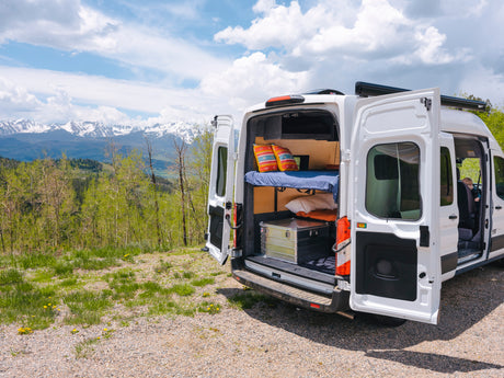 An RV rental, a white camper van with open back doors revealing a bunk bed and storage, parked before majestic snow mountains.