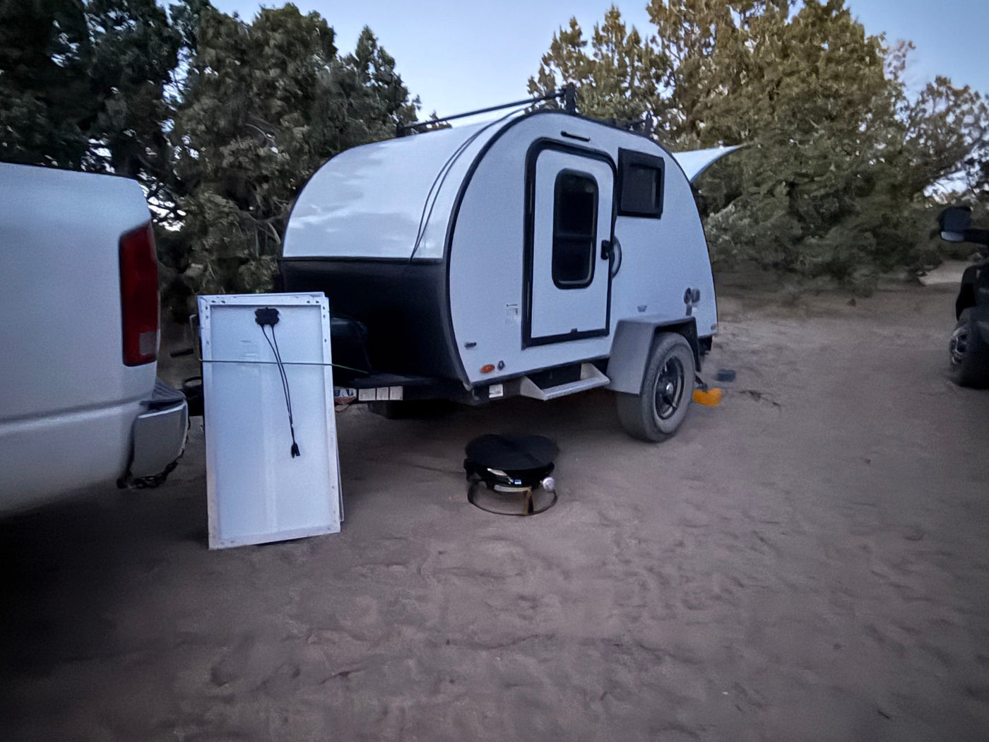 Teardrop RV rental for off-grid camping. Truck, solar panel, and grill set up on dirt amid trees. — photo 6