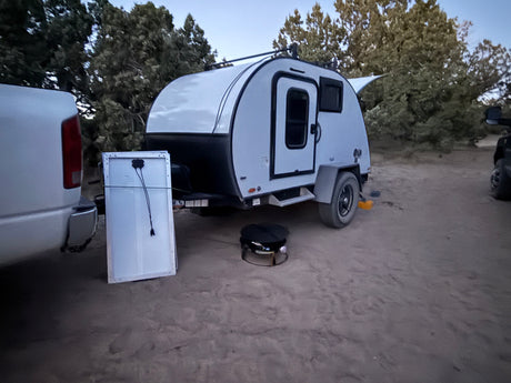 Teardrop RV rental for off-grid camping. Truck, solar panel, and grill set up on dirt amid trees.