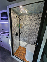 Modern RV rental interior featuring a compact shower with wood floor, toilet, and adjacent kitchenette.