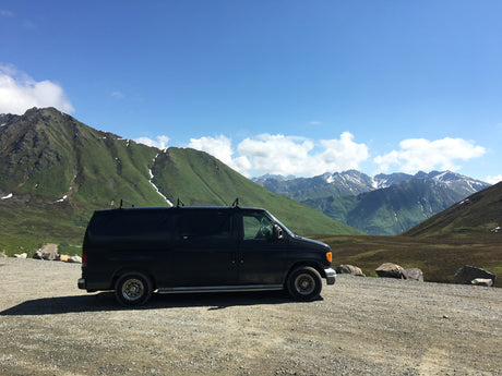 RV rental: Dark van on a gravel pullout amidst vast green, snow-dusted mountains under a clear blue sky. A rugged, scenic view.
